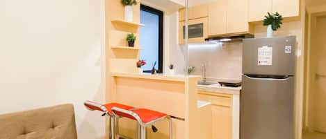 A compact kitchen with a stainless steel refrigerator, a microwave, and a sink. There are two bar stools and a small window above the sink.