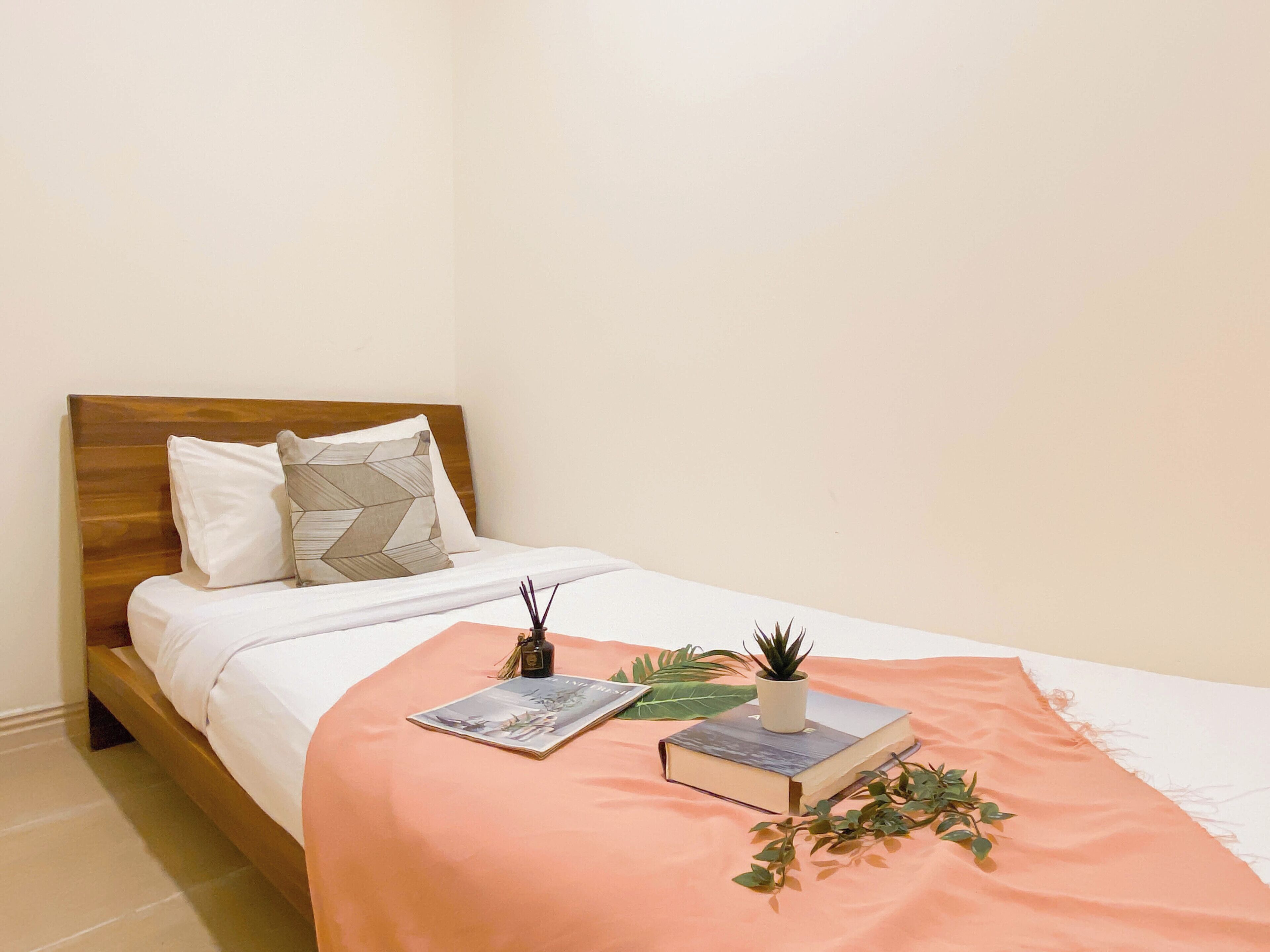 A neatly made bed with a wooden headboard, a white comforter, and a peach-colored blanket. There are two pillows, a book, a small potted plant, and a decorative item on the bed.