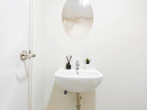 A white bathroom with a round mirror, a sink, and a small potted plant. - Warm Neutral Studio Room At Meikarta Apartment (Cikarang)