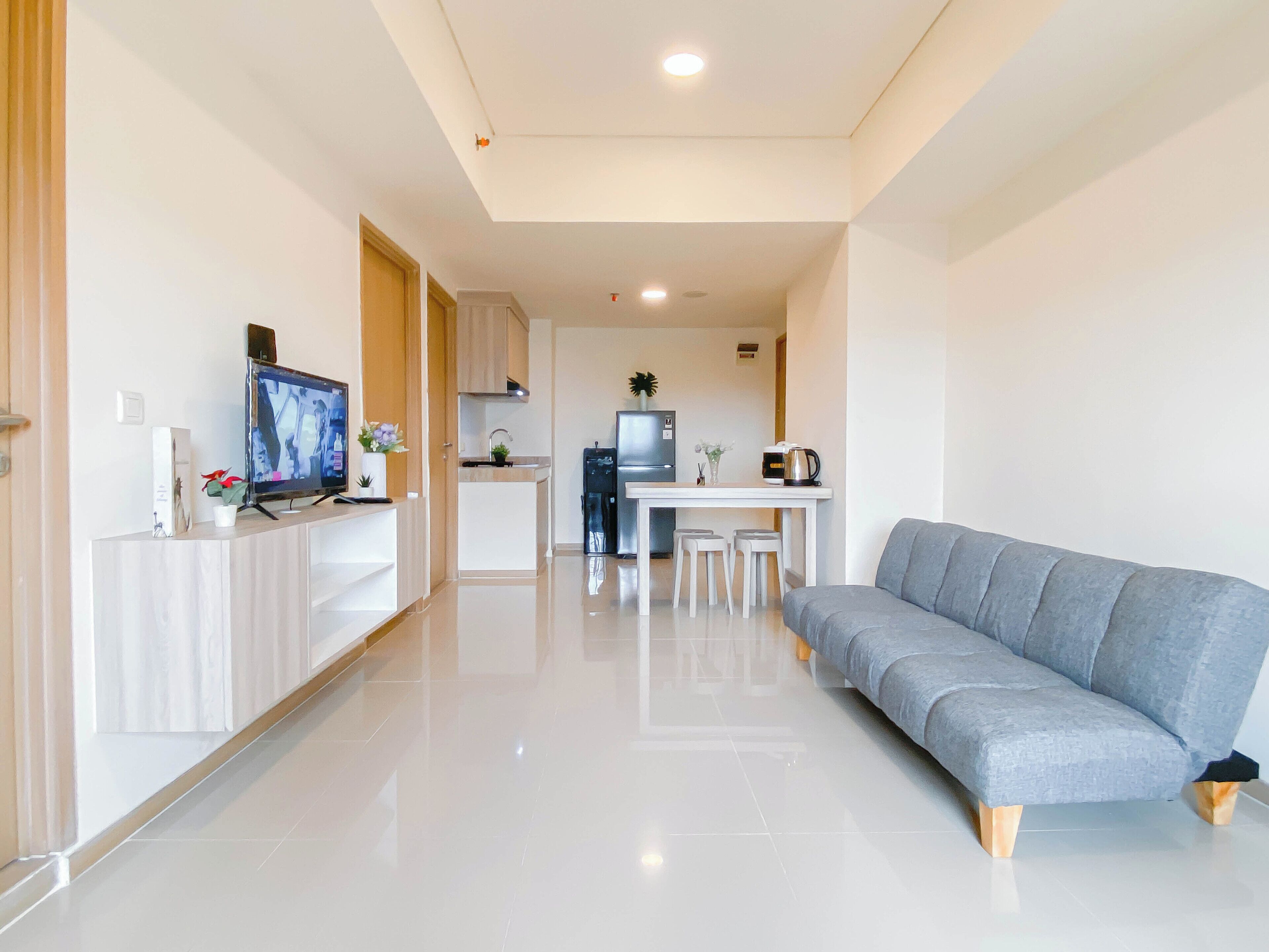 A modern living room with a grey sofa, a flat-screen TV, and a small kitchen area with a fridge, sink, and a coffee maker.