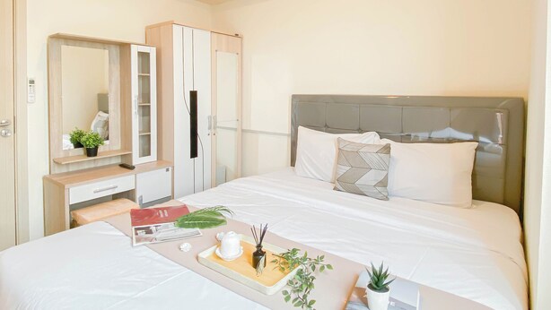 A hotel room with a bed, bedside table, dresser, and a large mirror. - Soft Scandinavian 3Br At Meikarta Apartment (Cikarang)