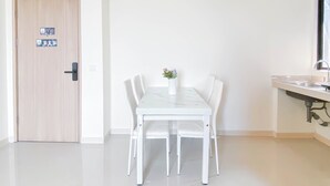 A modern, minimalist room with a wooden door, a white dining table with four chairs, a small vase with flowers, and a kitchen area with a sink and countertop. - Soft Scandinavian 3Br At Meikarta Apartment (Cikarang)