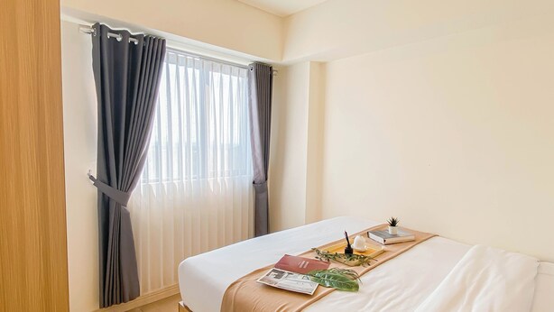 A hotel room with a bed, a window with curtains, a bedside table with a tray, and a small potted plant. - 3Br Apartment Near Dian Harapan School At Meikarta (Cikarang)