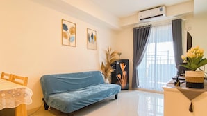 A living room with a blue sofa, a dining table with a lace tablecloth, a wall-mounted air conditioner, and a window with curtains. - 3Br Apartment Near Dian Harapan School At Meikarta (Cikarang)