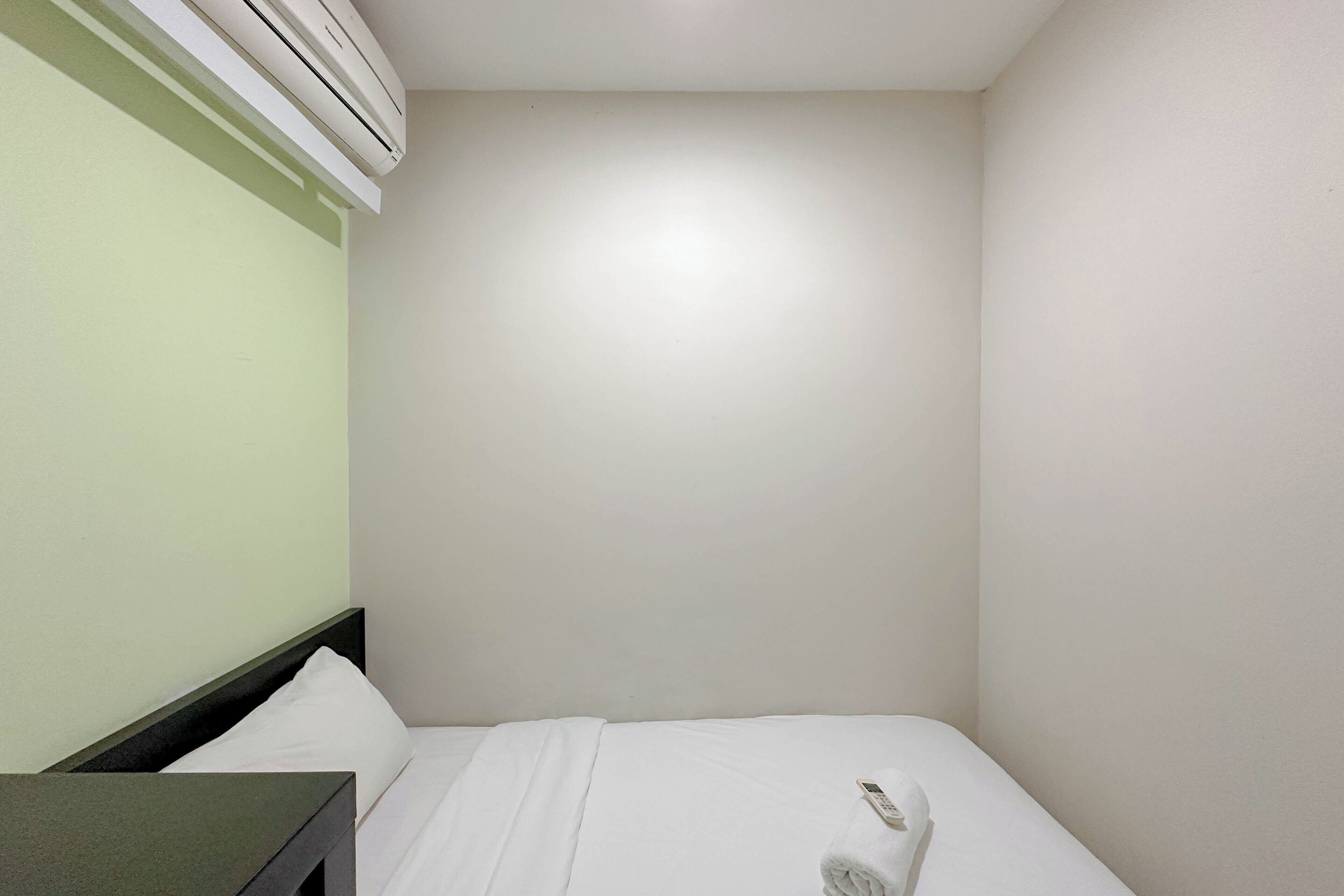 A small, clean room with a bed, a bedside table, and an air conditioning unit mounted on the wall.