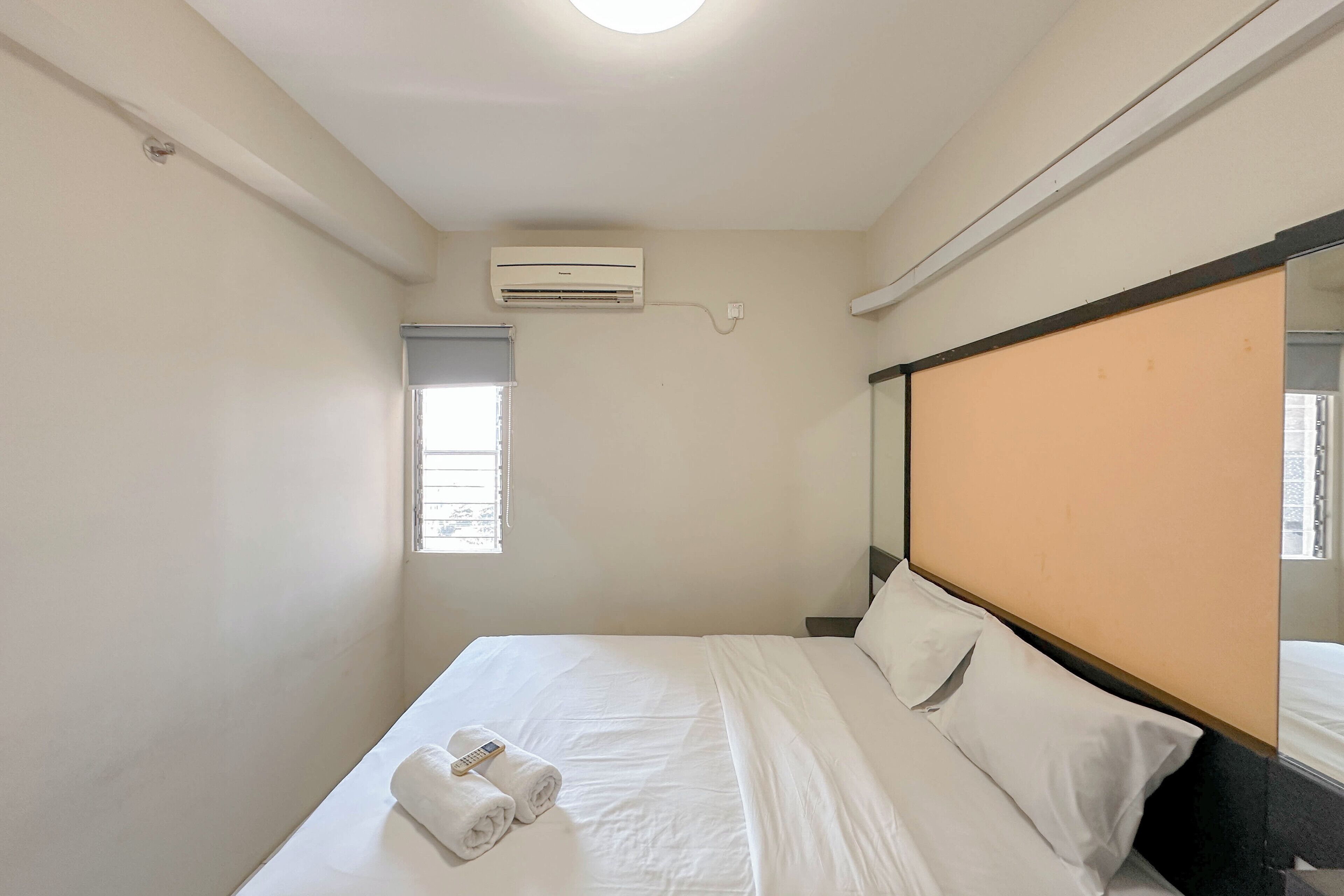 Une chambre d’hôtel avec un lit, du linge de lit blanc, une fenêtre avec des stores, un climatiseur et un miroir fixé au mur.