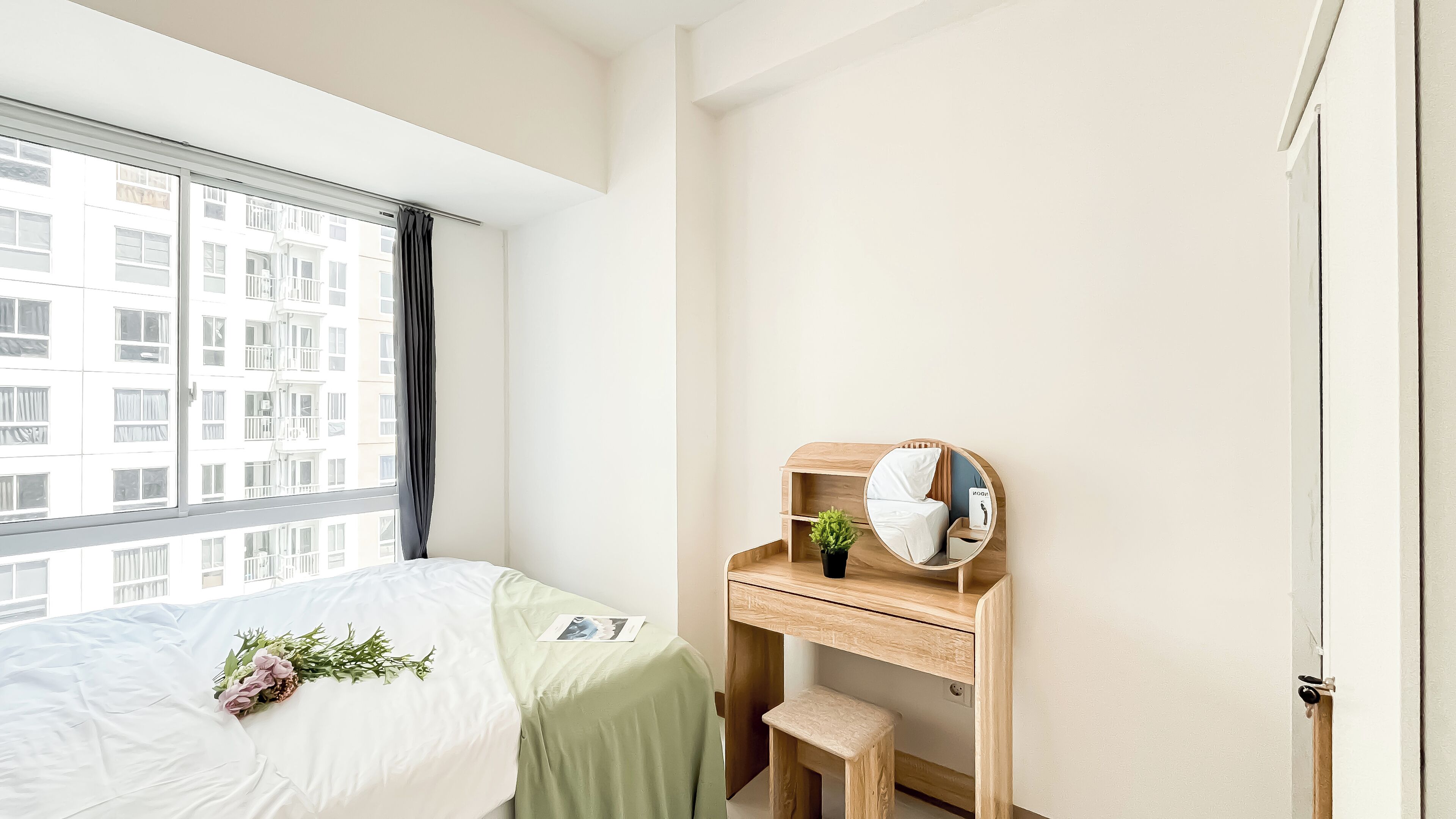 Een slaapkamer met een bed, een bureau, een spiegel en een raam met uitzicht op gebouwen.