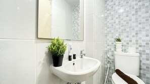 A modern bathroom with a white sink, mirror, and toilet. There is a potted plant on the sink counter and toiletries on the counter. - Soft Industrial Look Studio At Tokyo Riverside Pik 2 Apartment (Teluknaga)