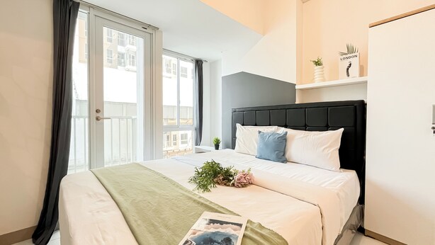 A hotel room with a bed, a window with curtains, a bedside plant, and a shelf with a sign reading "LONDON". - Soft Industrial Look Studio At Tokyo Riverside Pik 2 Apartment (Teluknaga)