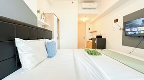 A modern hotel room with a bed, a flat-screen TV mounted on the wall, a kitchen area, and a door leading to another room. - Soft Industrial Look Studio At Tokyo Riverside Pik 2 Apartment (Teluknaga)