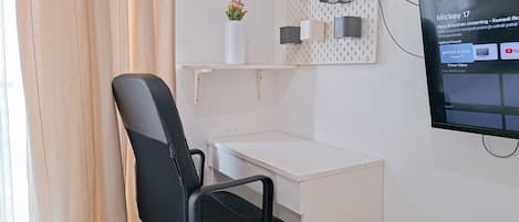 A hotel room with a black office chair, a white desk, a mounted television displaying a video player interface, and a wall-mounted shelf with a vase of flowers.