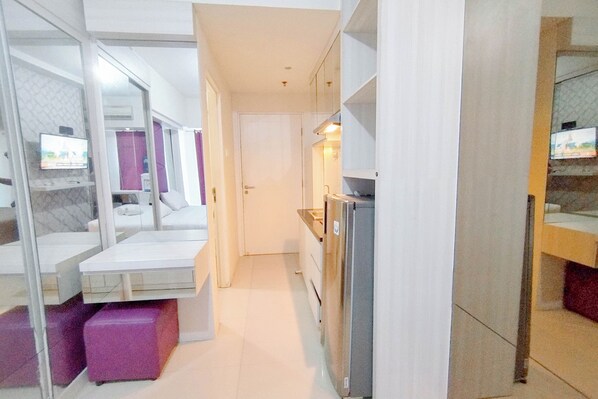 A modern hotel room with a glass partition separating the living area from the bedroom, a flat-screen TV mounted on the wall, and a small kitchenette with a refrigerator. - Purple Touch Studio At Orchard Supermall Mansion Apartment (Surabaya)