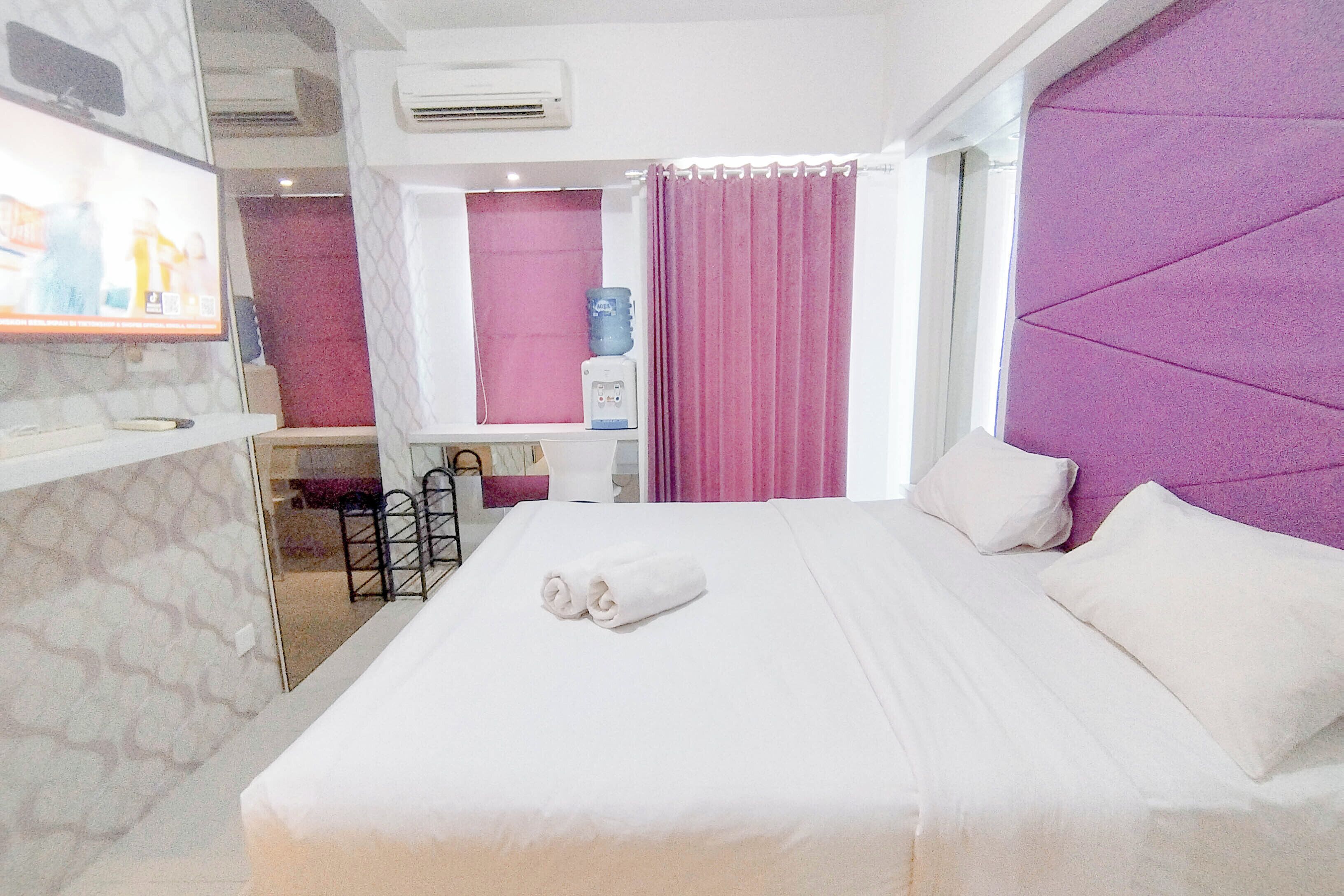 A hotel room with a bed, a TV, a water dispenser, and a bathroom visible through a glass partition.