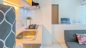 A modern kitchen with a stainless steel sink, a gas stove, and a small countertop. There is a potted plant on the counter and a vase with flowers. A sofa with a floral cushion is visible in the foreground. - Basic 2Br Apartment Podomoro Golf View (Gunung Putri)