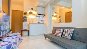 A modern living room with a grey sofa, colorful cushions, a coffee table, and a TV. A kitchen is visible in the background with white cabinets and a stove. - Basic 2Br Apartment Podomoro Golf View (Gunung Putri)