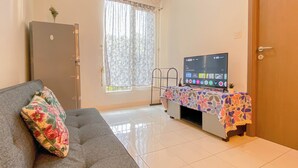A living room with a sofa, a television on a stand, a small table with a colorful tablecloth, and a window with curtains. - Basic 2Br Apartment Podomoro Golf View (Gunung Putri)