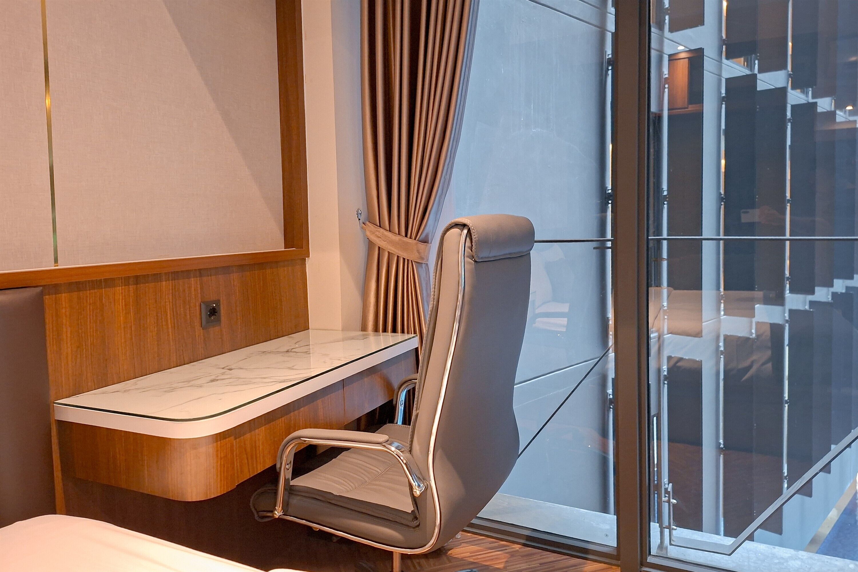 A modern hotel room with a glass partition, a cushioned chair, a small desk, and a view of a building through the glass.