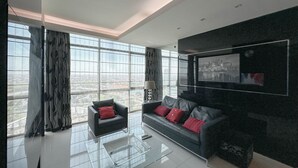 A modern living room with a glass coffee table, two sofas with red cushions, a large TV mounted on the wall, and large windows offering a city view. - Spacious And Comfortable Living 3Br Apartment At Aryaduta Residence (Surabaya)