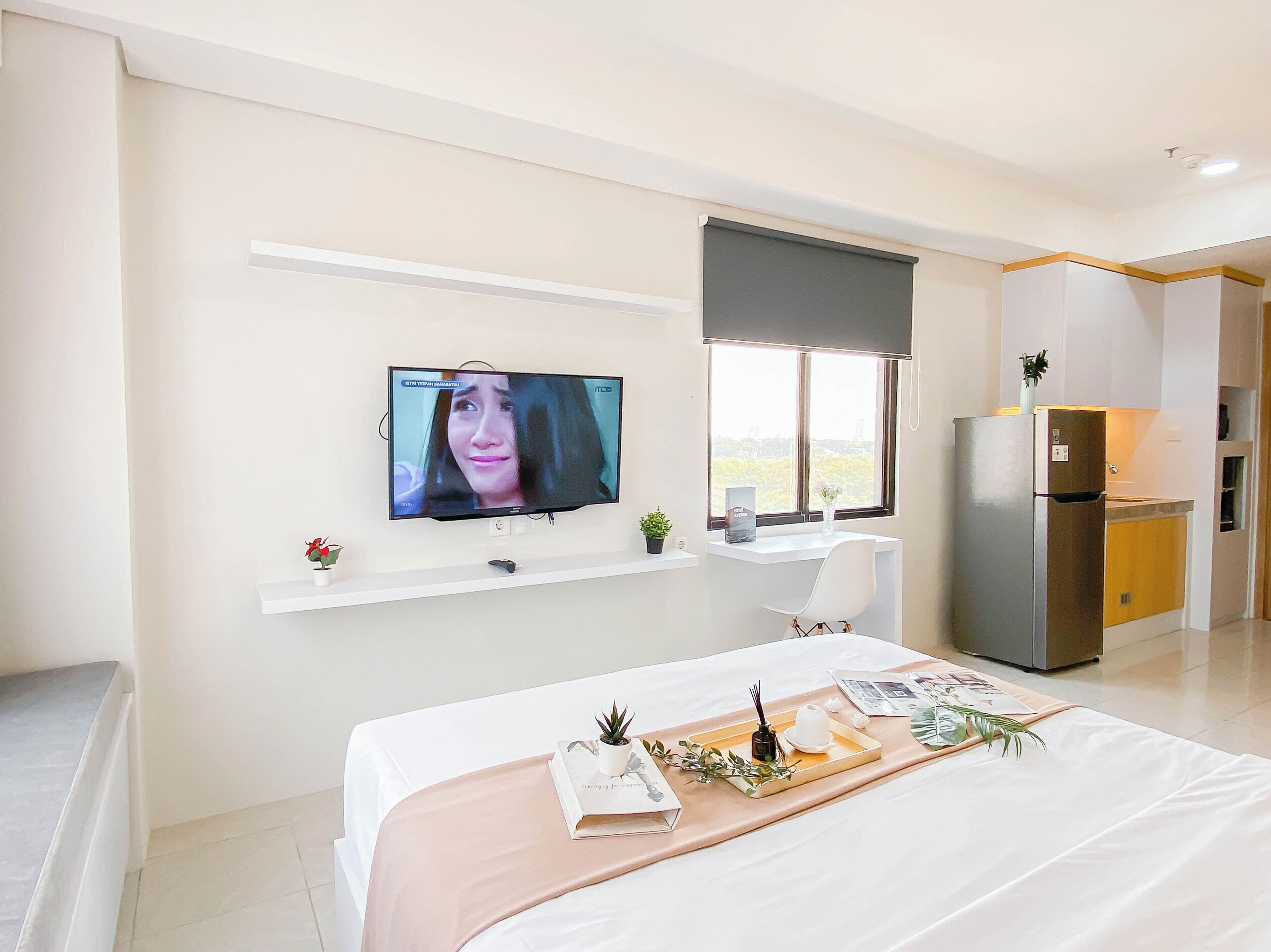 A modern hotel room with a bed, a TV mounted on the wall, a small desk with a chair, a window with a view, and a kitchenette.