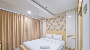 A neatly made bed with white linens and a folded towel, a wooden nightstand, a wall with a floral pattern, and a door with a gold frame. - Spacious 2Br At Green Park Yogyakarta Apartment (Depok)