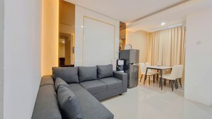 A modern living room with a grey sectional sofa, a small table with chairs, a refrigerator, and a coffee machine. - Spacious 2Br At Green Park Yogyakarta Apartment (Depok)