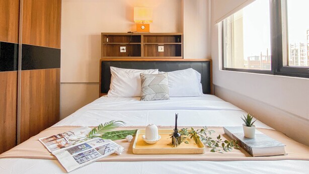 A neatly made bed with pillows, a bedside table with a tray of items, a nightstand with a lamp, and a window with a view of buildings. - Simple 1Br Near Dian Harapan School At Apartment Meikarta (Cikarang)