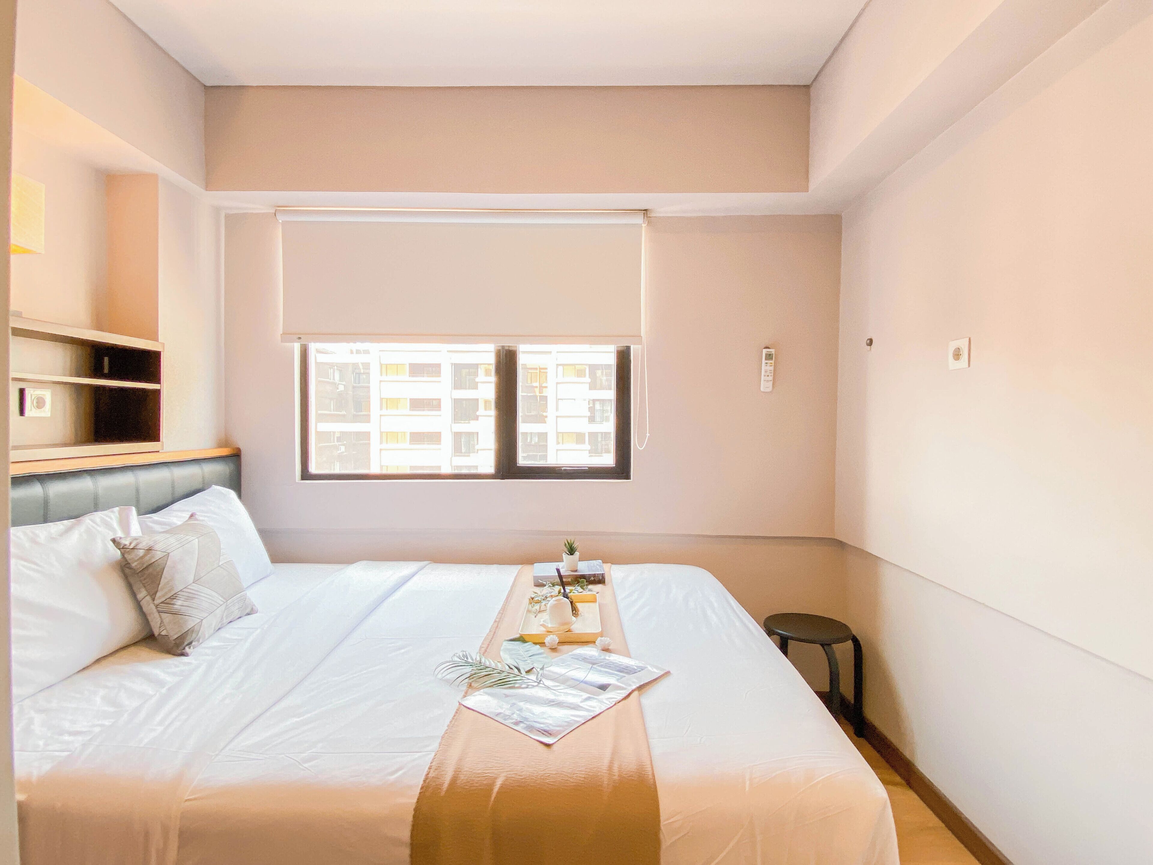 A modern hotel room with a bed, a small table with a tray, a window with blinds, and a bedside table with a lamp.