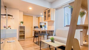 A modern kitchen with wooden cabinets, a dining table, and chairs. There is a sofa and a small table with a plant. - Simple 1Br Near Dian Harapan School At Apartment Meikarta (Cikarang)