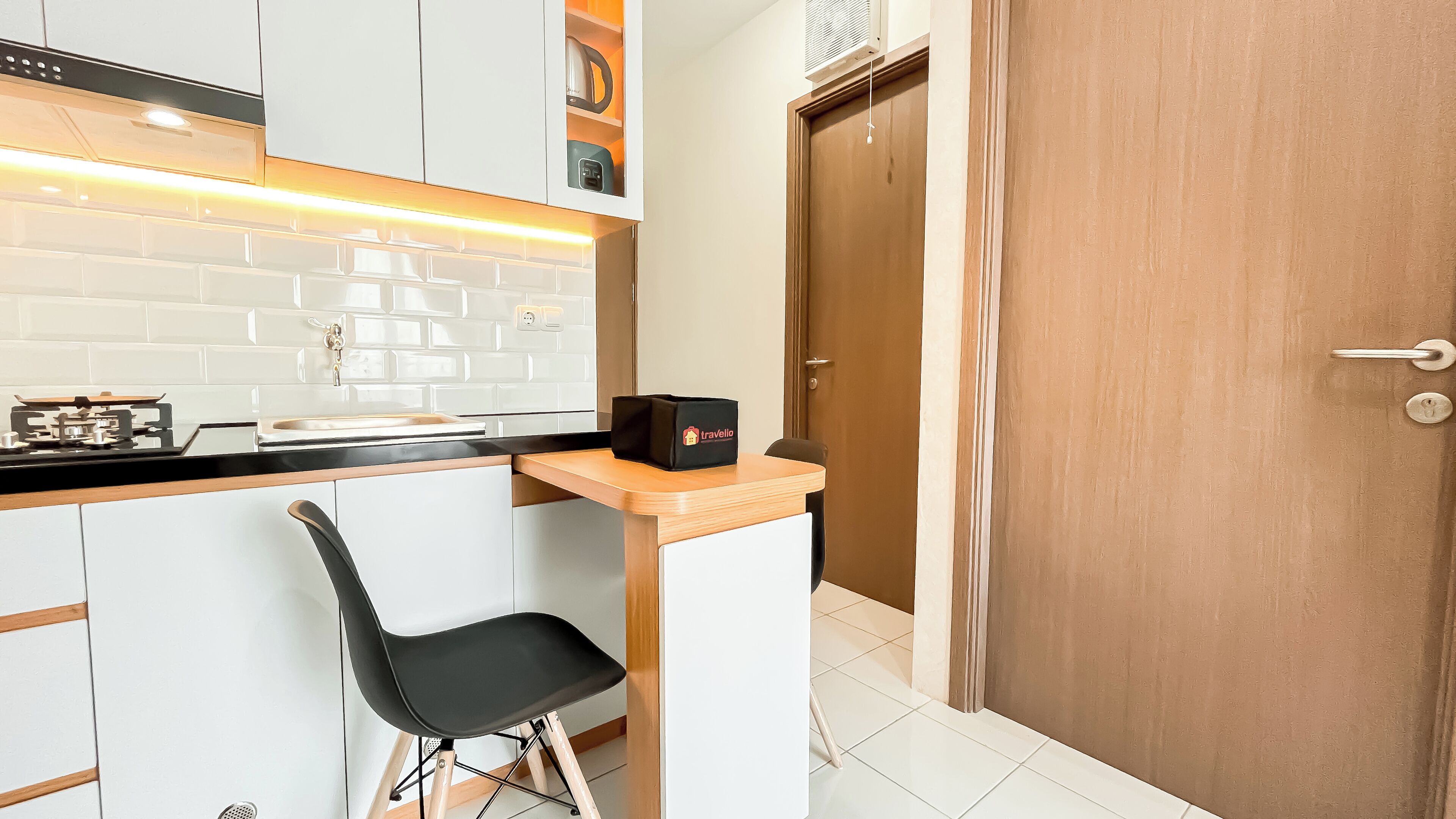 A compact kitchen with white cabinets, a black countertop, and a small black chair. There's a wooden door to the right and a black bag labeled 'Travelia' on the countertop.