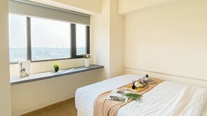 Egy hálószoba, nagy ablakkal, bézs színű ágytakaróval borított ággyal, és egy kis polccal, melyen növények és egy szobor található.