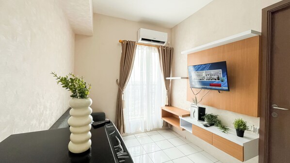 A modern hotel room with a black desk, a white vase with green plants, a mounted TV, and a wooden shelf with a small plant. - Minimalist 2Br Apartment At Podomoro Golf View (Gunung Putri)