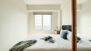 A bedroom with a bed, a nightstand, a window with blinds, and a view of the city. - Minimalist 2Br Apartment At Podomoro Golf View (Gunung Putri)