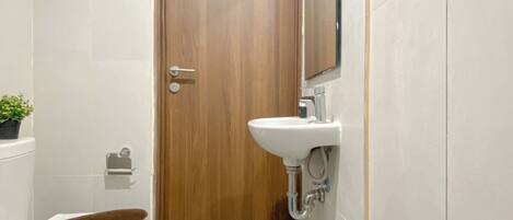 Un baño con puerta de madera, inodoro blanco, lavabo con espejo encima y una pequeña planta en maceta a la izquierda.