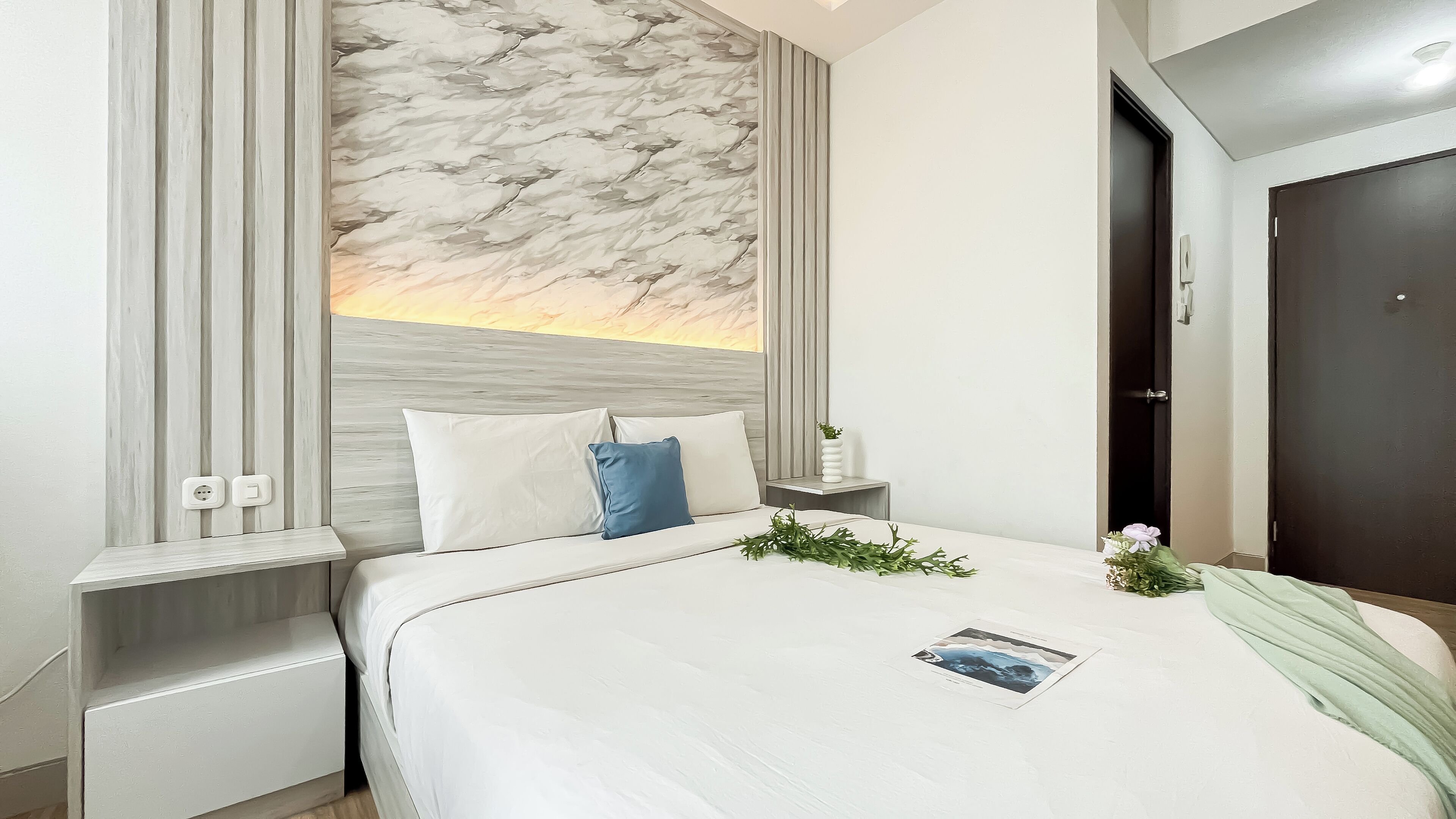 A modern hotel room with a large bed, a nightstand, a wall-mounted power outlet, and a door.