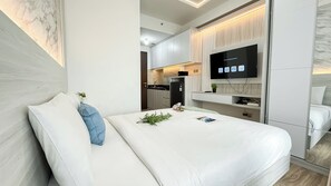 A modern hotel room with a large bed, a flat-screen TV mounted on the wall, a small kitchen area, and a mirror reflecting the room. - Modern Studio At Transpark Cibubur Apartment (Depok)