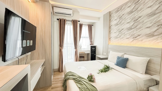A modern hotel room with a flat-screen TV, a desk with a chair, a bed with pillows, and a marble-patterned wall. - Modern Studio At Transpark Cibubur Apartment (Depok)