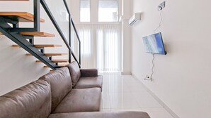 A modern living room with a staircase, a flat-screen TV mounted on the wall, and a large window with sheer curtains.