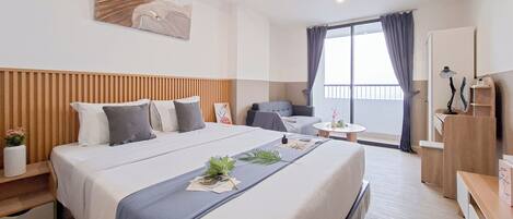 A hotel room with a large bed, a desk, a chair, a small table, and a balcony with curtains.