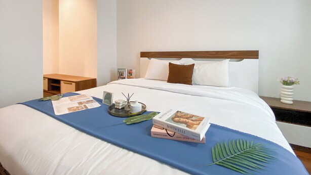 A neatly made bed with a blue bedspread, a wooden headboard, and a bedside table with a lamp. - Homey 2Br Apartment At Lucky Tower Residence (Jakarta)