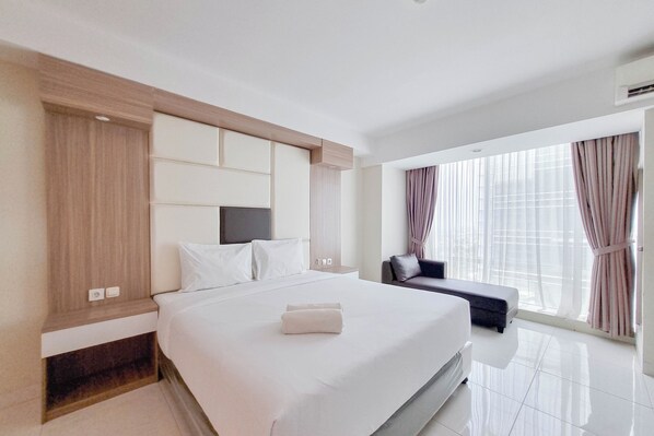A modern hotel room with a large bed, a flat-screen TV, a bedside table, and a large window with curtains. - Tidy Studio At Mataram City Apartment (Ngaglik)