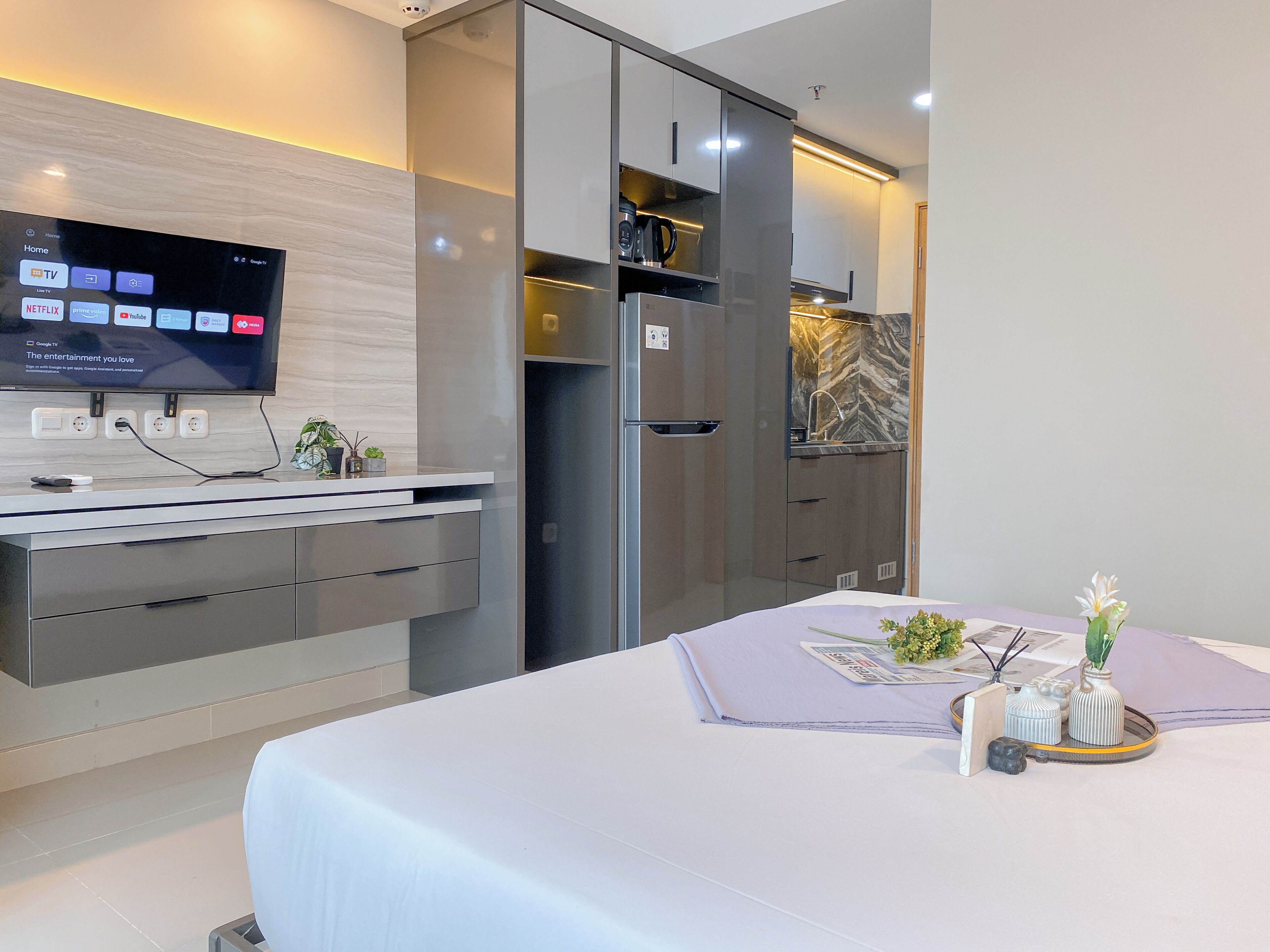 A modern hotel room with a flat-screen TV mounted on the wall, a built-in kitchen with a refrigerator and microwave, and a dining area with a table set for two.