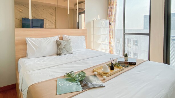 Camera d'albergo con un letto grande, una testiera in legno, uno specchio, una finestra con tende e un vassoio con un libro e una pianta.