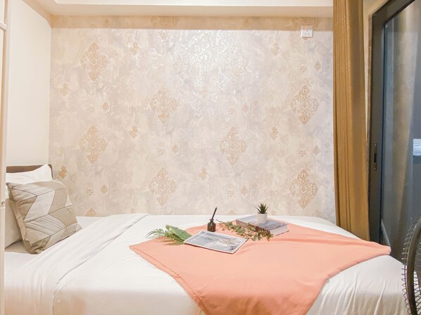 A neatly made bed with a peach-colored blanket, a small potted plant, and a magazine on the bed. - Earthtone 2Br At Meikarta Apartment (Cikarang)