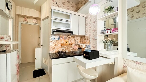 A compact kitchen with white cabinets, a black countertop, a gas stove, and a small sink. There is a white bar stool and a decorative mirror above the sink. - Compact 2Br Apartment Near Unj At Green Pramuka City (Jakarta)