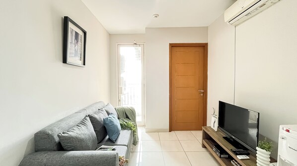 A living room with a grey sofa, a flat-screen TV on a wooden stand, and a door leading to another room. - Scandinavian Minimalist 2Br At Cinere Bellevue Suites Apartment (Depok)