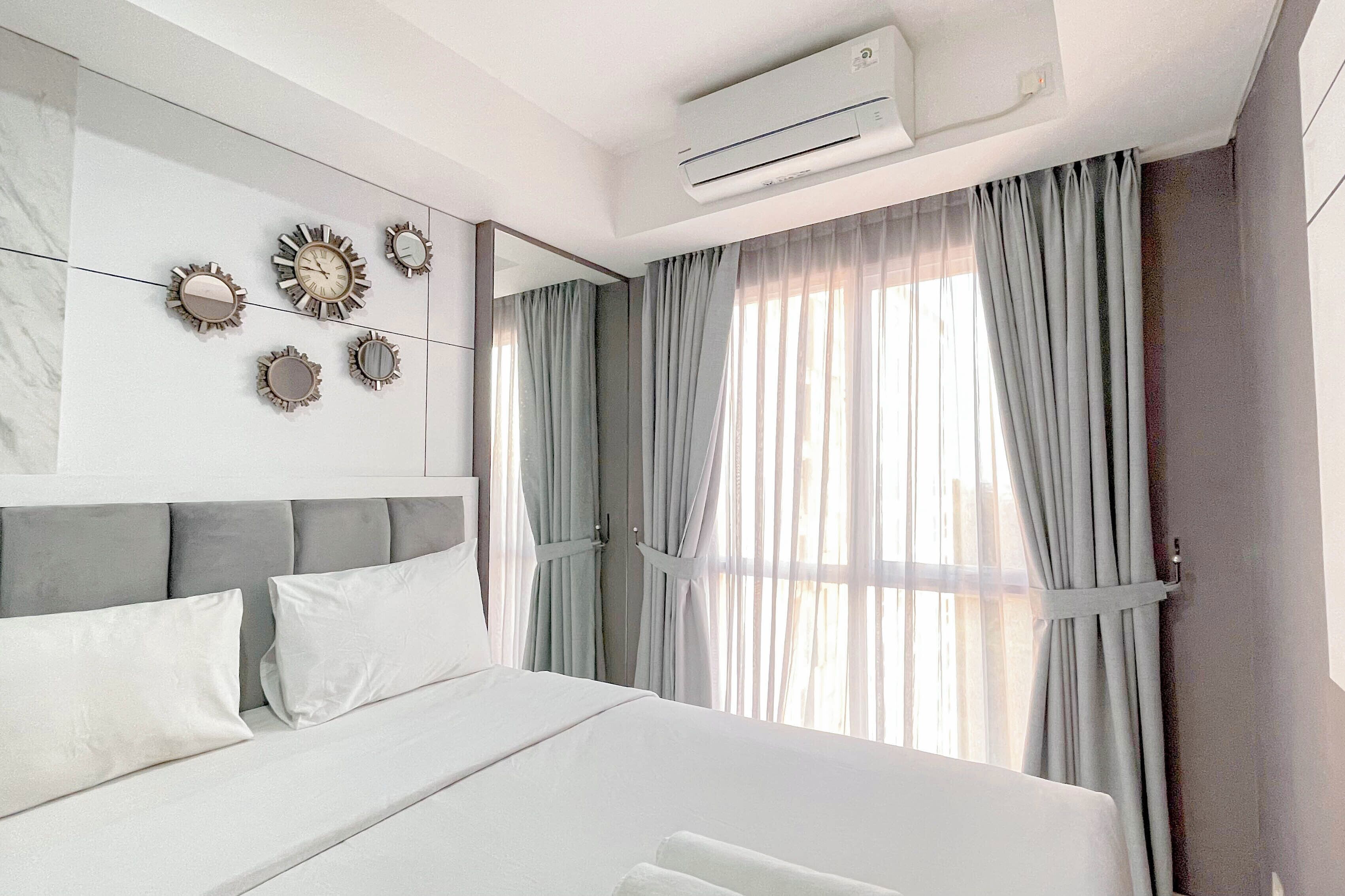 A modern hotel room with a large bed, a wall-mounted air conditioner, and a window with light curtains.