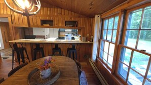 A wooden interior with a kitchen area, a dining table with chairs, and a large window.