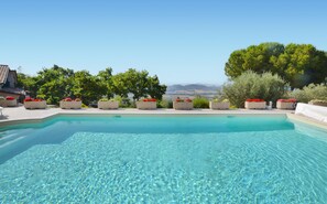 A clear swimming pool with a view of distant hills, surrounded by planters with red flowers, and a building with a tiled roof in the background. - Amazing apartment in Magione (Magione)