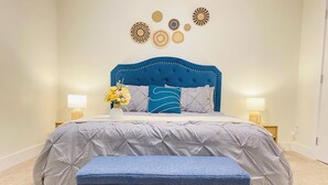 Une chambre à coucher avec une tête de lit capitonnée, une couvre-lit grise, un banc bleu et des œuvres d’art décoratives accrochées au mur.
