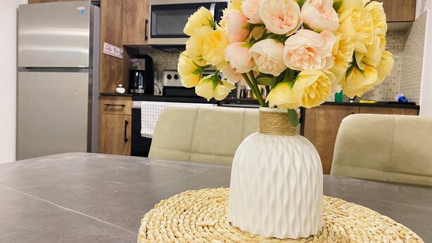 Une table à manger avec un set de table en fibres tressées, un vase blanc orné de fleurs et, au fond, une cuisine.